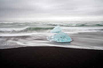 Crystal clear and blue ice chunks washes up on the black lava sand by the waves on diamond beach in Jokulsarlon glacier lagoon. 