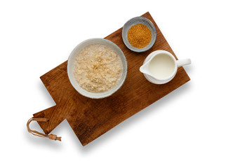 A nutritious bowl of porridge, a small bowl of brown sugar and a jug of milk, on a wooden board, isolated on a white background with a drop shadow.