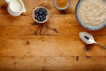 A bowl of nutritious porridge, a small bowl of blueberries, a jug of milk and bowl of brown sugar, with a spoon on a wooden background, with room for copy.