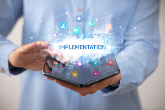 Businessman holding a foldable smartphone with IMPLEMENTATION inscription, technology concept