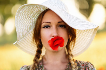 Portrait of beautiful redhead woman in white hat on green field with poppy flower in her mouth