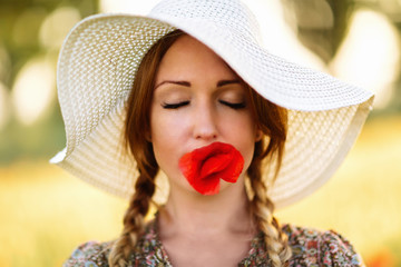 Portrait of beautiful redhead woman in white hat on green field with poppy flower in her mouth