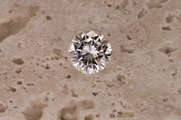 A colorless round cut diamond sits isolated on a light rough stone background.