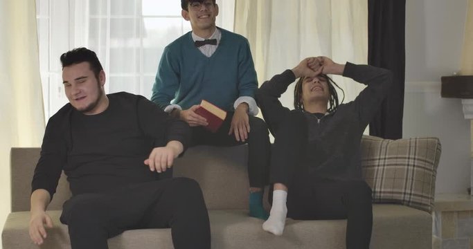 Portrait of three Caucasian adult students watching sports match on TV. Football fans gesturing emotionally as their male friend reading book at the background. Cinema 4k ProRes HQ.