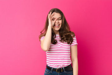 young pretty woman smiling happily with funny face, joking and looking through peephole, spying on...