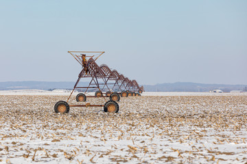 Rusty, old center pivot irrigation system sits idle in harvested cornfield covered in snow during winter. Maintenance, repair, and upkeep concept