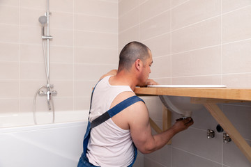 Closeup male plumber worker in blue denim uniform, overalls, fixing sink in bathroom with tile...