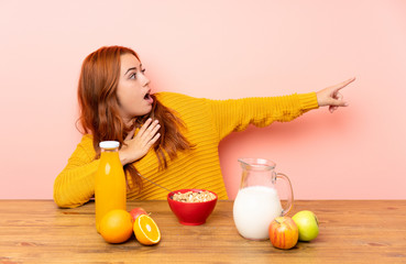 Teenager redhead girl having breakfast in a table pointing finger to the side