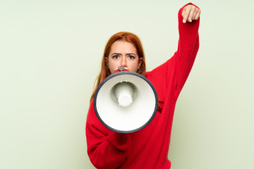 Teenager redhead girl with sweater over isolated green background shouting through a megaphone