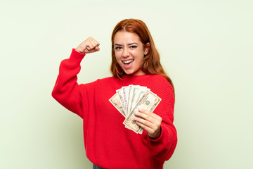 Teenager redhead girl with sweater over isolated green background taking a lot of money