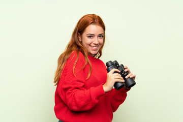 Teenager redhead girl with sweater over isolated green background with black binoculars