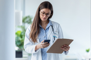 Pretty female doctor using her mobile phone while reviewing medical reports in medical consultation.