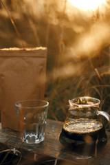 Glass kettle with coffee, cup and brown pack with coffee on background of rural countryside herbs in sunset. Alternative coffee brewing outdoors. Atmospheric rustic moment. Vertical image