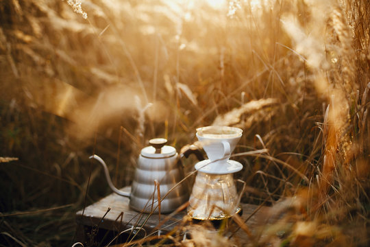Steel kettle and glass flask dripper with filter on background of rural countryside herbs in sunset. Alternative coffee brewing outdoors. Atmospheric rustic moment. Space for text