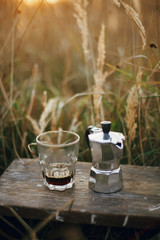 Geyser coffee maker and glass cup with fresh coffee in sunny warm light in rural countryside herbs. Atmospheric rustic moment. Alternative coffee brewing in travel. Vertical image