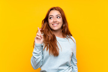 Teenager redhead girl over isolated yellow background with fingers crossing and wishing the best