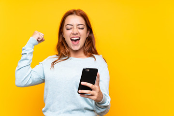 Teenager redhead girl over isolated yellow background with phone in victory position