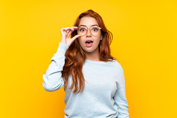 Teenager redhead girl over isolated yellow background with glasses and surprised