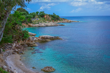 Naklejka premium Beautiful landscape – sea coast: turquoise calm water, stones and rocks on the beach, cloudy sky and green trees. Corfu Island, Greece, Kassiopi village