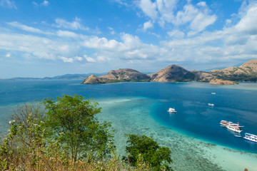 View from top of a cliff on Kelor Island, Komodo, Indonesia. Island is surrounded with white sand beaches and turquoise water. There is another island in the back.There are boats anchored to a shore.