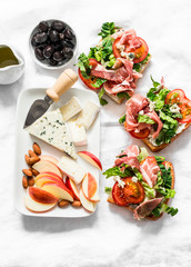 Appetizer, aperitif, snack table - gorgonzola, brie cheese, dried olives, apple, almonds and prosciutto, tomatoes, green salad sandwiches on a light background, top view