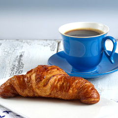 Cup and croissant on a white plate is on the table of gray boards.