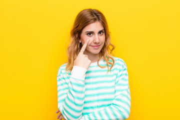 young pretty blonde woman keeping an eye on you, not trusting, watching and staying alert and vigilant against yellow wall - obrazy, fototapety, plakaty