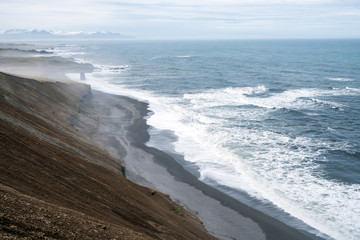 Steep cliffside with volcanic beach and waves crashing to shore. Mountains with snow in the background. From the southcoast of Iceland and the atlantic ocean. View towards Vestrahorn and Hofn.
