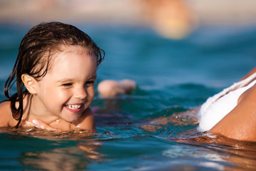 Young blond mother in blue bikini standing in water and helping to swim her small smiling daughter in sunshine on summer day. Family vacations and travelling concept