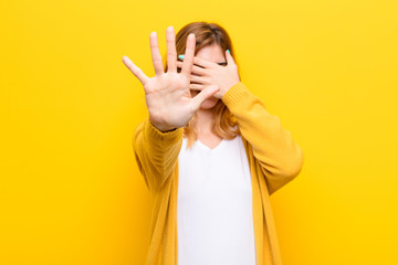 young pretty blonde woman covering face with hand and putting other hand up front to stop camera,...