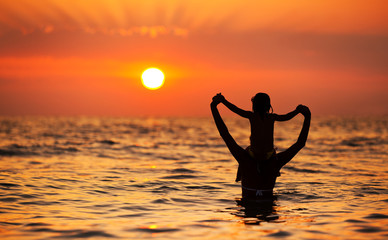 Young mother in white bikini standing in water and playing with her small daughter at golden sunset...