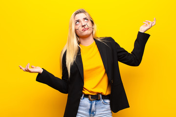 young pretty blonde woman shrugging with a dumb, crazy, confused, puzzled expression, feeling...