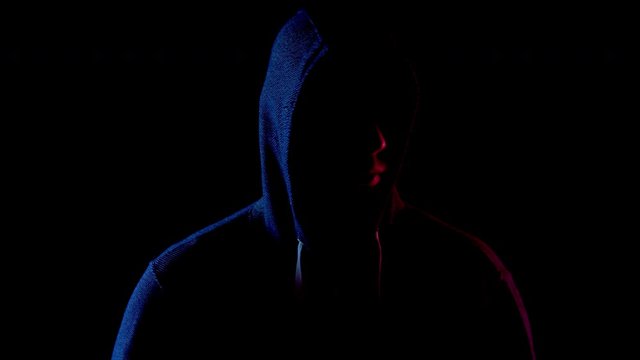 A young man in a hood looks around. An attacker is standing in the dark. Blue and red light falls on a person on a black background.