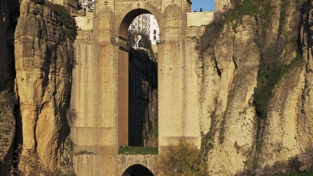 Tilt down shot of stone bridge in Ronda city cliffs and small waterfall under. Water stream under Ronda bridge and cliffs, evening sun