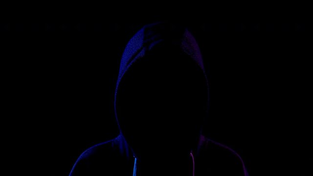 A young woman is standing in the hood hiding her face. An attacker is standing in the dark. Blue and red light falls on the person on a black background.