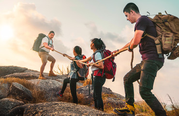 Young asian four hikers climbing up on the peak of mountain. People helping each other hike up a mountain at sunlight. Giving a helping hand. Climbing. Helps and team work concept - obrazy, fototapety, plakaty