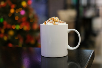 White mug with cocoa or coffee with cream with caramel syrup in a cafe on the background of a Christmas tree bokeh garlands. Cappuccino or lat with topping in a cup.