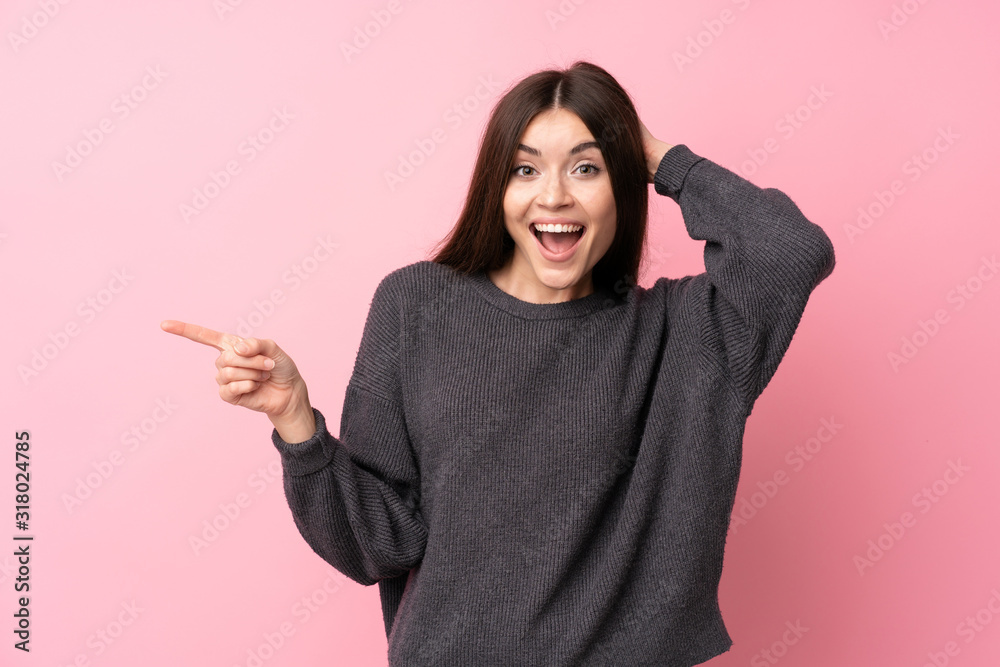 Wall mural Young woman over isolated pink background surprised and pointing finger to the side