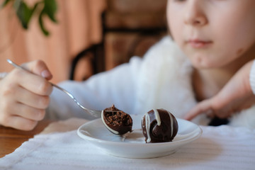 A little girl in white clothes sits at a table by the window. eating a fork with a white saucer dessert. chocolate balls made of white and black chocolate