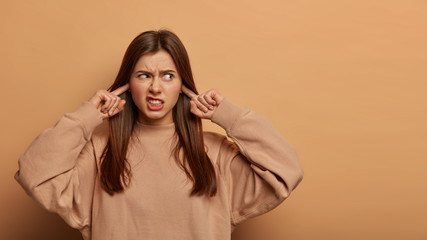 Horizontal view of young irritated woman uwills hearing bothering noise, plugs ears and frowns face from anger, annoyed by loud music, wears sweatshirt, stands over beige wall. Turn volume, please!