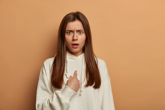 Excuse me. Puzzled indignant woman points at herself, frowns discontentedly face, being confused in doing something wrong, asks question, wears snow white sweatshirt, poses over beige studio wall