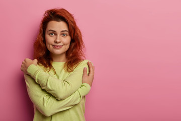 Tender romantic ginger woman hugs herself, embraces own body, touches shoulders gently, has pleasant smile, wears green comfortable sweater, stands against pink background. Love and care concept