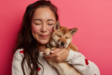 Close up shot of pleased Asian woman carries pedigree dog closely near face, closes eyes from...
