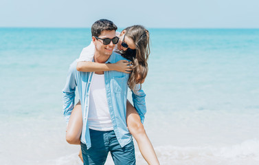 Handsome man giving piggy back to his girlfriend walking on beach behind blue sky or honeymoon trip. Beach couple laughing in love romance on travel honeymoon vacation summer holidays romance.