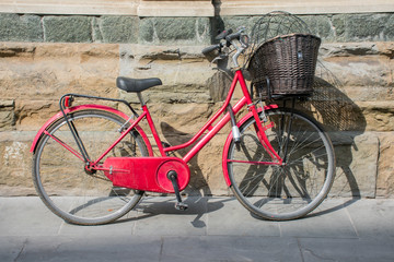 Fototapeta na wymiar red bicycle parked throwing its shadow on the sidewalk of an urban street. Nobody in the scene uses the vehicle. The bicycle has a wooden brown basket at front handlebar. Horizontal photo