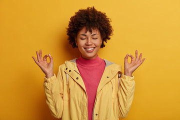 Concentrated cheerful curly woman keeps both hands in okay gesture, meditates indoor, has eyes...