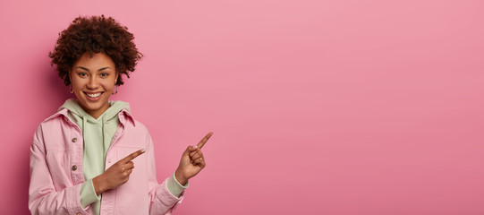 Studio shot of joyful positive woman wears sweatshirt and denim pink jacket, points index fingers right, smiles happily, advertises blank space for your text information or promotion on rosy wall