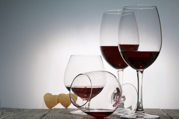 Glasses with red dry wine. One of them lies on its side with the remains of wine. Nearby are hearts cut out of paper. Stand on wooden boards. Shot in backlight.