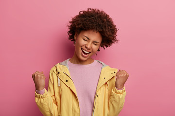 Obraz na płótnie Canvas Carefree black woman clenches fists with joy, tilts head and feels like winner, celebrates victory, closes eyes with pleasure, wears casual jumper and jacket, laughs out happily, isolated on pink wall