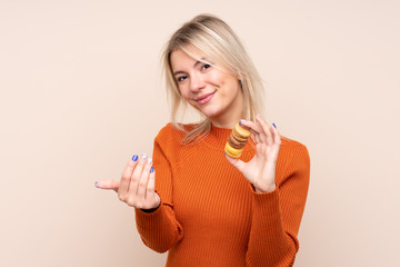 Young blonde Russian woman over isolated background holding colorful French macarons and inviting to come